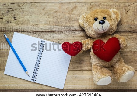 A photo of Teddy bear holding a heart-shaped pillow on wood table with notepad paper