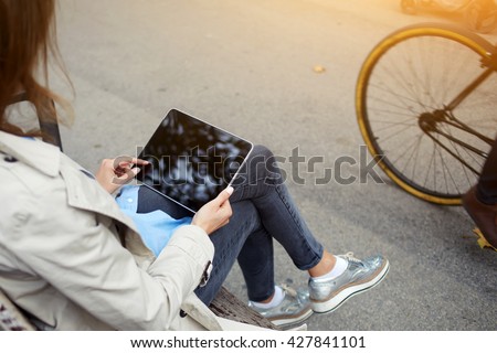 Back view of young hipster girl is making purchase at the on-line store via touch pad, while is sitting on the street in spring day.Closely image of woman is holding digital tablet with mock up screen