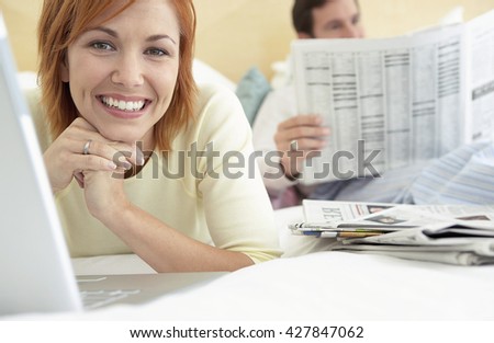 Young woman using a laptop smiling at the camera with her husnband reading newspaper in the background