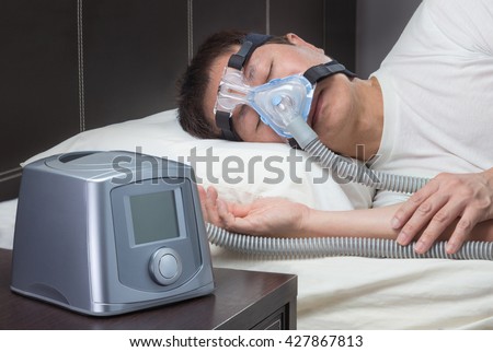 Asian man with sleep apnea using CPAP machine, wearing headgear mask connecting to air tube Royalty-Free Stock Photo #427867813