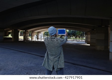 Modern young man with tablet pc touch taking photos under a bridge with wool cap