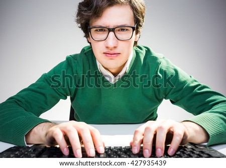 Serious young man in green pullover and glasses sitting at desk, keyboarding and looking at the camera 