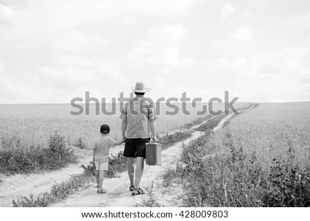 Back view black white photography dad holding his little son by hand walking away along empty outdoors country field road on summer sunny day