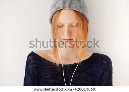Cute student girl with ginger hair and freckles wearing gray hipster cap looking down while listening to audiobook with earphones, preparing for exams at university standing against white background 