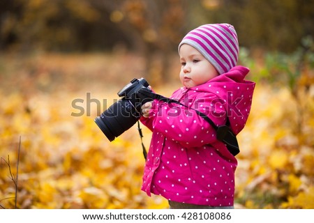 A small girl is photographer, kid holds a camera and takes photo of autumn landscape, child stands in the autumn park and girl is dressed in warm hat and jacket. Royalty-Free Stock Photo #428108086