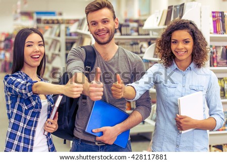 Attractive girls and guy are holding books, showing Ok sign, looking at camera and smiling while standing at the bookshop