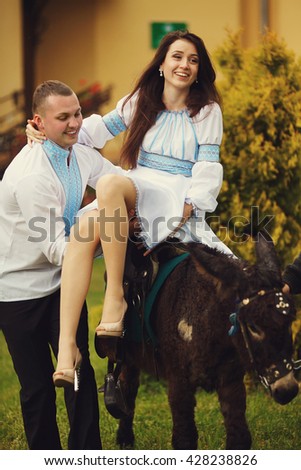 A picture of a pretty brunette sitting on the donkey and a man in embroidered shirt holding her