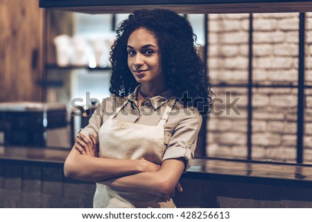 Waiting for the first customer. Young cheerful African woman in apron keeping arms crossed and looking at camera with smile while standing at bar counter