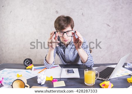 Crazy exhausted businessman sitting at messy office desk and looking at the camera through eyeglasses