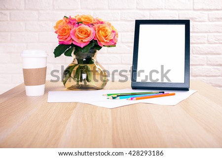 Front view of wooden desktop with roses, coffee cup, blank picture frame and colorful pencils on white brick wall background. Mock up
