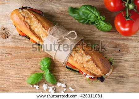 Papered vegetarian baguette submarine sandwich with grilled eggplant, pepper and feta cheese on wooden chopping board over dark background. Top view Royalty-Free Stock Photo #428311081