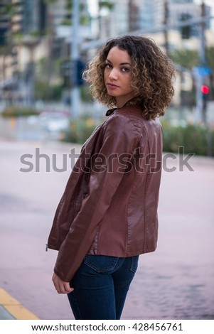 Young slim female in leather jacket and jeans on a sidewalk, looking behind her