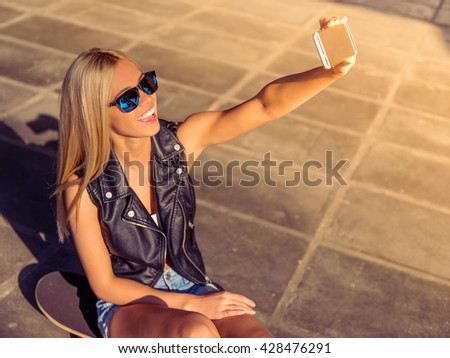 Beautiful blonde girl in glasses is making selfie using a smart phone and smiling while sitting on her skateboard in skate park