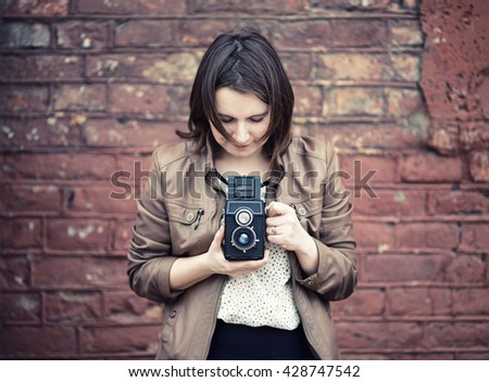 Pretty young woman holding retro camera and taking photo on vintage brick wall background. Selective focus on camera. Toned photo with copy space. Vintage style photo.