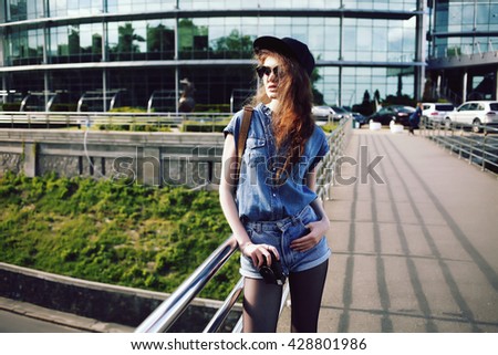 Funny cheerful hipster girl backpacker smiling and taking photo with vintage retro camera, wearing swag hat, sunglasses and denim shorts and shirt, amazing view. Image toned style instagram filters