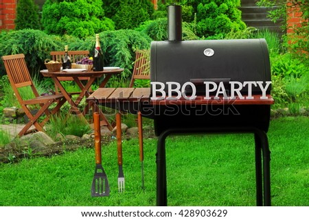 Summer Weekend BBQ Scene With Charcoal Grill On The Backyard Lawn And Outdoor Wooden Furniture In The Background