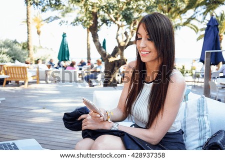Young latin female is writing an e-mail while sitting in the lounge bar. Smiley girl is holding the smart phone while having rest in a street cafe.