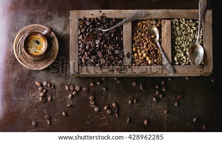 Green and brown decaf unroasted and black roasted coffee beans in old wooden box, and ceramic cup of fresh making coffee over dark brown textured background. Banner top view Royalty-Free Stock Photo #428962285