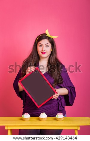 Close-up plump plus size brunette woman with curly bright makeup and pink lips with the tablet for the text in the hands.  On a pink background vintage cement wall. Unusual emotion