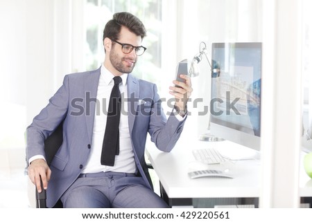 Shot of a handsome businessman using mobile phone while sitting at office in front of computer. 