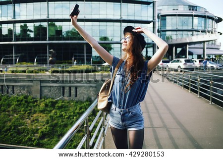 Funny cheerful hipster girl backpacker smiling and taking photo with vintage retro camera, wearing swag hat, sunglasses and denim shorts and shirt, amazing view. Image toned style instagram filters