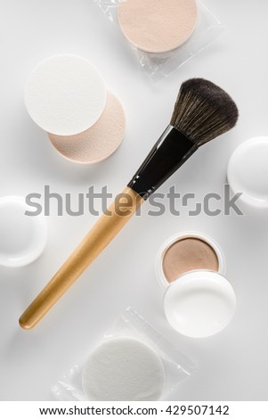 The brush with the cosmetics for face in the center of the picture. White jars with brown and flesh concealers and powder with special sponges for putting on the foundation.