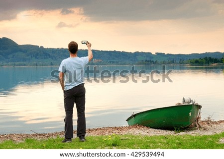 Man in blue shirt take photo by smart phone. Silhouette at fishing paddle boat at mountains lake coast.
