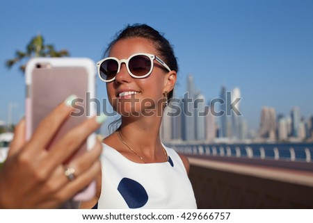 Tourist woman taking mobile phone selfie picture at Palm Jumeirah, Dubai. Beautiful girl holding smartphone for self-portrait photo with view of Dubai skyscrapers during summer travel vacation.