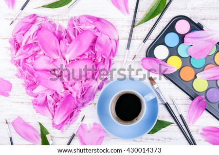 Workspace with cup of coffee and flowers. Watercolor, paintbrush and petals of peonies on white wooden background. Top view