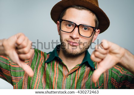 hipster man showing a sign of dislike, thumbs down, wears glasses and hat, isolated on gray background