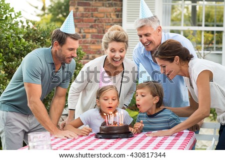 Happy generation family watching girl blowing out birthday candles at picnic table outside. Happy little girl celebrating birthday with family. Brother helping sister blow candles on her birthday.