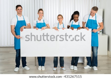 Group Of Happy Cleaners In Uniform Holding Blank Banner In Office