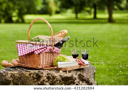 Elegant picnic with red wine, fresh grapes, cheese, baguette and sausages arranged on a rustic stone table in a lush green spring or summer park, with copy space