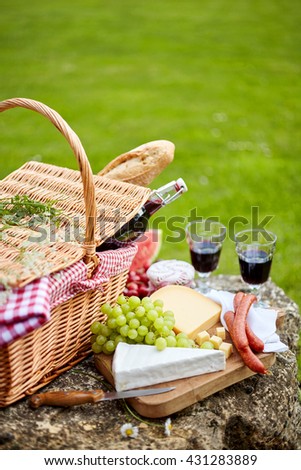 Stylish picnic with red wine, fruit and cheese served with spicy sausages and fresh baguette on a cutting board balanced on a rock in a green lawn with copy space