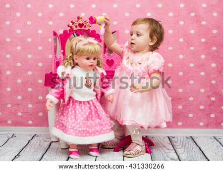 little girl on a pink background, plays with a doll posing and fancies. Princess fashionably dressed, painted, preening and combing and dress her her doll at the mirror