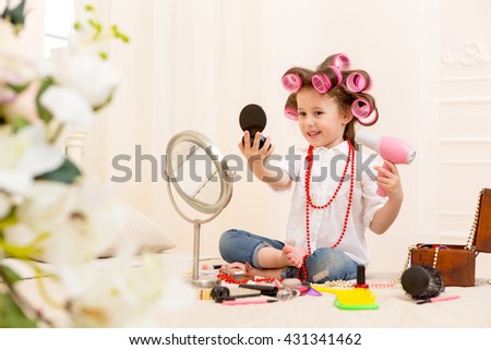 
Happy child. Portrait of a pretty little girl looking in a small mirror and holding a pink hairdryer on a bed in the bedroom . Girl laughing with pink hair curlers on her head Royalty-Free Stock Photo #431341462