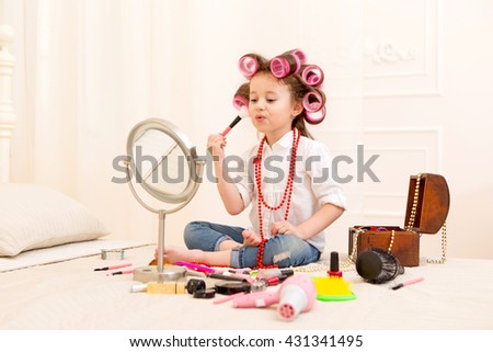 Happy child. Little pretty girl makes a make-up on a bed in the bedroom . Girl laughing with curlers on her head , and brushes for makeup in the hands Royalty-Free Stock Photo #431341495