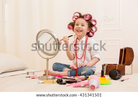 Happy child. Little pretty girl makes a make-up on a bed in the bedroom . Girl laughing with curlers on her head , and brushes for makeup in the hands Royalty-Free Stock Photo #431341501
