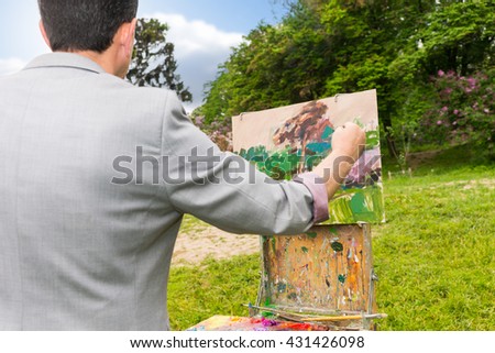 Over the shoulder view of a middle-aged fashionable male  painter working on a sketchbook painting a garden scene with flowers outdoors