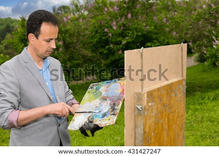 Pensive fashionable middle-aged artist holding a palette standing in front of a sketchbook during an art class in a forest