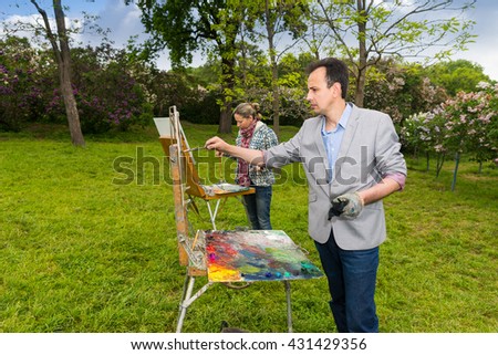 Two modest  painters standing in front of their sketchbooks painting pictures with  oils and acrylics paint during an art class in a park