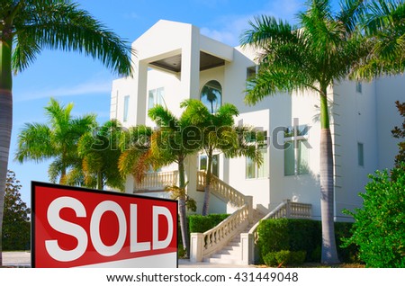 Beautiful purchased luxury residential home with front stairways and lush green palm trees against a blue morning sky with a bright SOLD sign in the front yard.