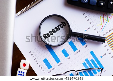 Magnifying glass on "Research" text on chart, dice, spectacles, pen, laptop calculator on wooden table - business, banking, finance and investment concept