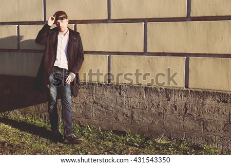 Gorgeous man in a raincoat standing in front of a brick wall. Blond. In black glasses.  Romantic image. Oldshool 70s colour graded.