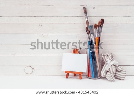 White background with brushes in the bank, a small easel and a teddy bear. Space for text.