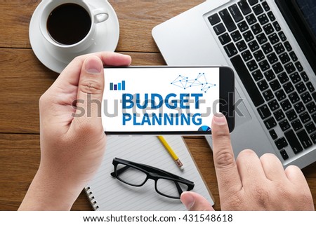 BUDGET PLANNING message on hand holding to touch a phone, top view, table computer coffee and book