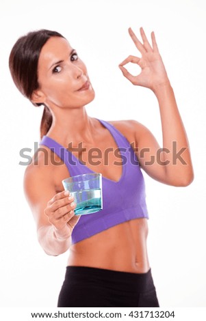 Healthy brunette woman offering a glass of water, gesturing perfect sign, looking at camera licking her lips while wearing violet and black gymnastic clothing, isolated