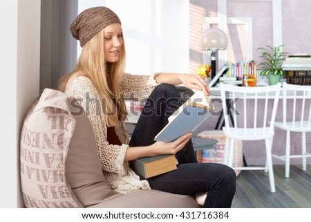 Blonde female student reading book at home, wearing cap.