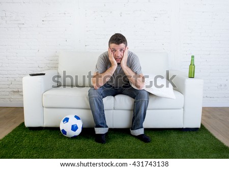 crazy football fan cheering watching television soccer match suffering stress nervous and excited biting his fingernails sitting on sofa couch with grass carpet and ball emulating stadium pitch