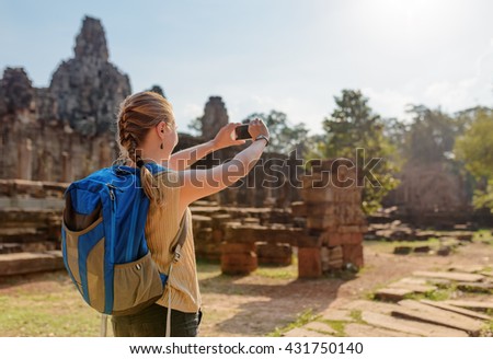 Young female tourist with blue backpack and smartphone taking picture among mysterious ruins of ancient Bayon temple in Angkor Thom. Siem Reap, Cambodia.